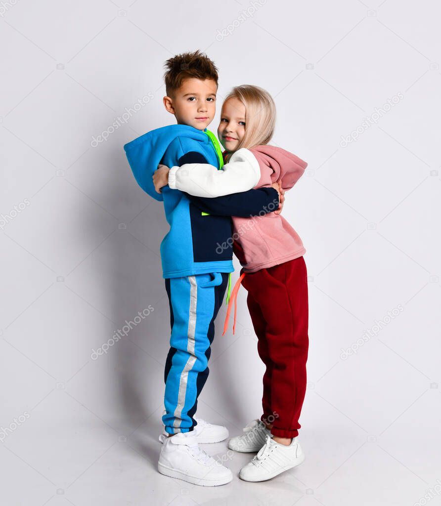 Little kids, brother and sister, in hoods, colorful tracksuits and sneakers. They holding hands, posing sideways isolated on white