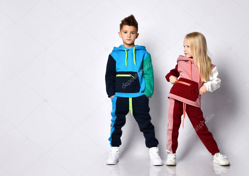 Little kids, boy and girl, in colorful tracksuits and sneakers. They posing isolated on white studio background. Hands in pockets