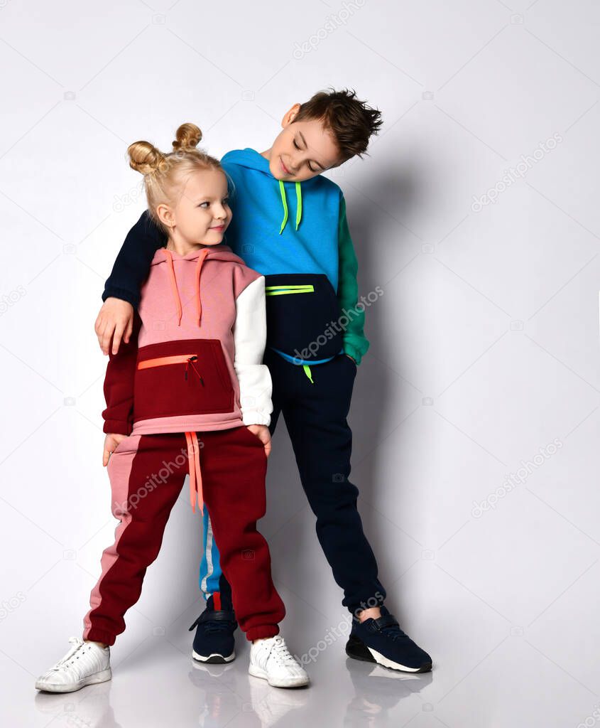 Smiling elder brother putting hand on shoulder of younger sister. Both kids in stylish track suits with cute hairdos. Portrait isolated on light grey