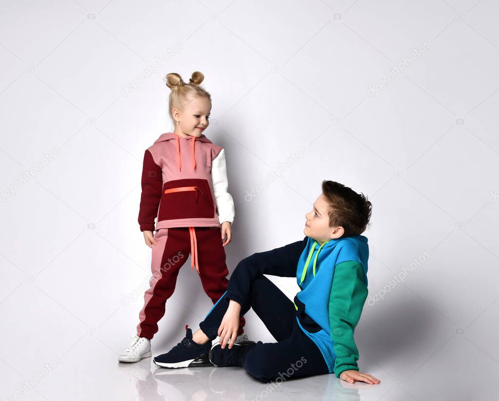 Children, brother and sister, in colorful tracksuits and sneakers. They looking at each other, smiling, posing isolated on white
