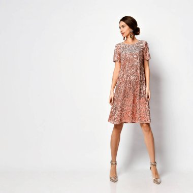 Zarif saç stili ve kalın küpeli genç bir kadın. Parlak, kısa, pembe bir elbise ve beyaz tenli gümüş ayakkabıları var.