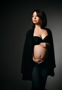 Siyah şallı, pantolonlu ve sütyenli, çıplak karnı olan esmer bir kadın. Yan bakıyor, gri stüdyo arka planında poz veriyor.