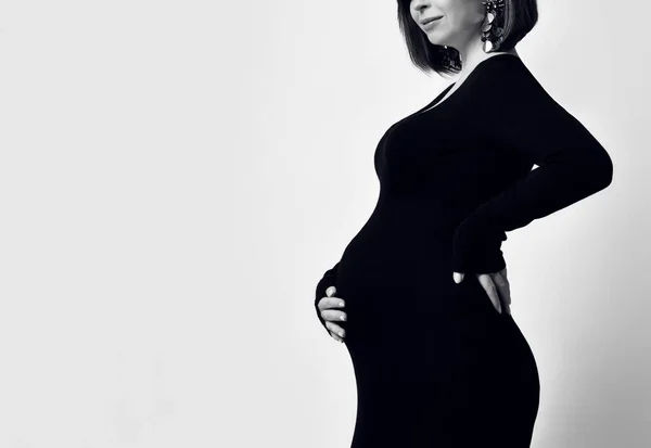 검은 색 꽉 끼는 드레스에 귀걸이를 한 임신 한 여성. 그녀는 그녀의 배에 손을 얹고 옆으로 포즈를 취했다. 흑백 사진. — 스톡 사진