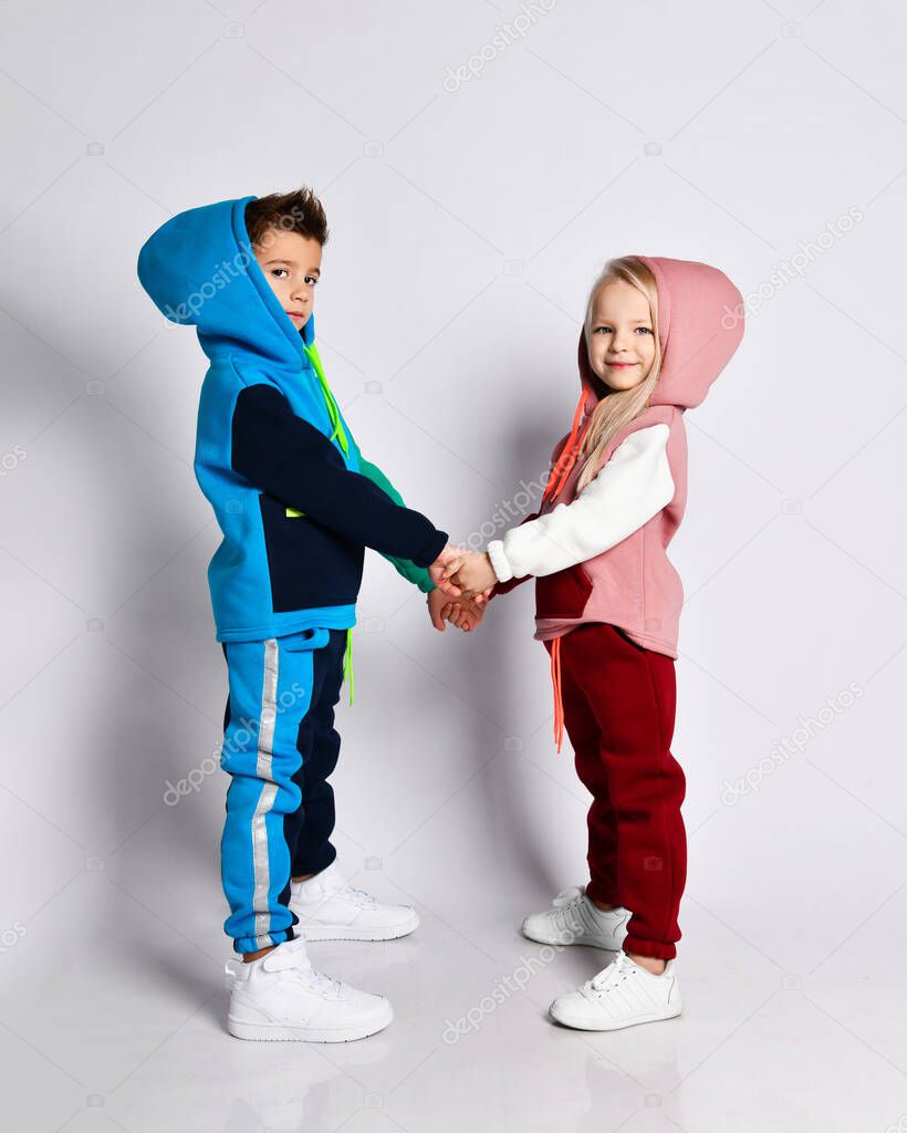Little kids, brother and sister, in hoods, colorful tracksuits and sneakers. They holding hands, posing sideways isolated on white