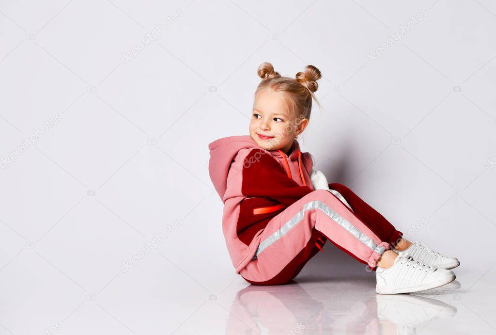 Little blonde girl with buns hairstyle, in colorful tracksuit, sneakers. She laughing, sitting on floor, posing isolated on white