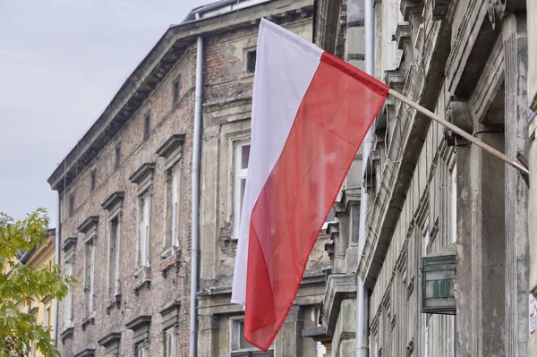 Η πολωνική κόκκινη και άσπρη σημαία κρέμεται στην πρόσοψη ενός παλιού σπιτιού σε μια συννεφιασμένη μέρα ενάντια σε έναν γκρίζο ουρανό. Εθνική εορτή σε χώρα ή πόλη, ημέρα σημαίας ή ημέρα ανεξαρτησίας στην Κρακοβία, Πολωνία. — Φωτογραφία Αρχείου