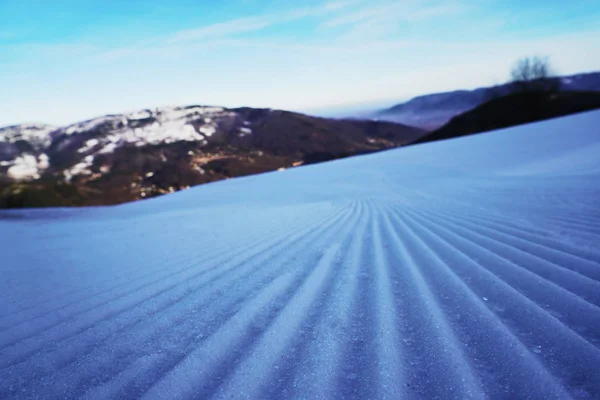 Pista de esqui de neve branca após snowcat ou tratamento ratrak no fundo de montanhas cobertas de neve e céu azul com nuvens. vista do solo, foco seletivo. esquiar e snowboard descanso ativo, esporte . — Fotografia de Stock