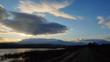 Gün batımında dağ gölü boyunca yürümek ya da bisiklete binmek için yol. Suyun üzerinde bulutlar ve ışık dalgaları, Zywiec 'te evleri ve ağaçları olan tepeler.