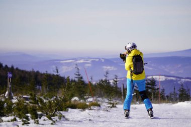 Parlak mavi ve sarı kayak elbiseli bir kadın dağın zirvesinde güneşli ve açık bir günde dağların manzarasını fotoğraflıyor. Aktif kış tatili, dağlarda kayak ve snowboard..