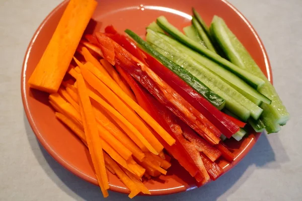 Bright seasonal vegetables cut into strips lie on a plate. Healthy natural food, fresh snack. Vegan and vegetarian, RAW. Green cucumber, red pepper and orange carrot for further cooking. — Stock Photo, Image