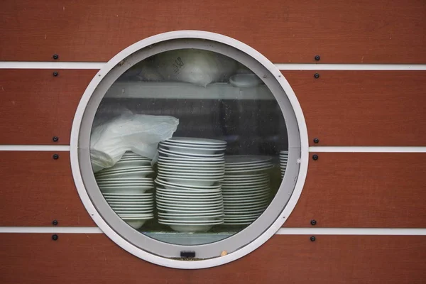 Un gran ojo de buey redondo en el lado marrón de la nave, a través del cual se pueden ver platos apilados y platos. restaurante cocina en el barco, comida rápida —  Fotos de Stock