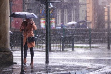 Şemsiyenin altında kahverengi paltolu bir kadın yoğun bir yağmurda yoğun bir caddeyi geçiyor. Şehirde şiddetli yağış var. Şehir sokaklarını sel bastı