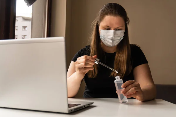 A girl in a medical mask works during the quarantine at a computer with a test tube in her hands. Prevention of coronavirus. Remote work at home during pandemic