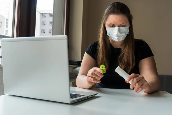 A girl in a medical mask works during the quarantine at a computer with a test tube in her hands. Prevention of coronavirus. Remote work at home during pandemic