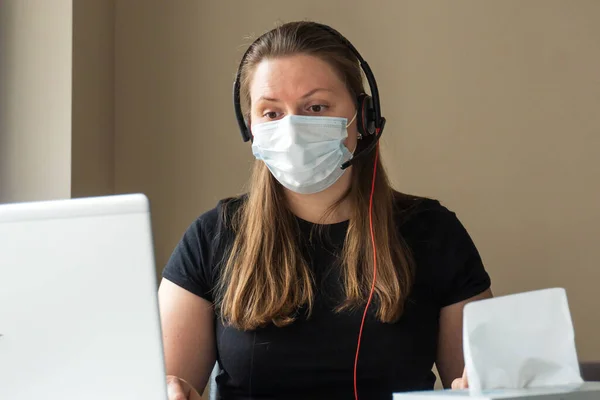 A girl in a medical mask works at home at the computer during the quarantine due to the coronavirus. Prevention of respiratory diseases. Remote work at home.