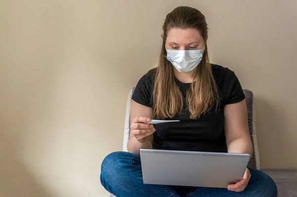 A girl in a medical mask holds a thermometer in her hand, which shows a high value of body temperature. Symptoms of coronavirus. Remote work at home during pandemic. Prevention of respiratory diseases.