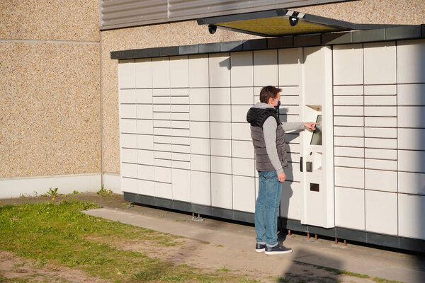 Молодой человек в синей маске забирает посылку из "Коробок" за посылками. бесконтактная или бесконтактная доставка товаров и продуктов во время эпидемии коронавируса или пандемии.
