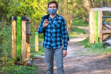 Tıbbi maske takan Avrupalı genç bir adam koronavirüs salgını sırasında Park 'ta ya da ormanda yürüyor. İnsanların sosyal mesafesi. Bahar ceketi giymişti.