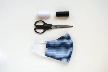 Koronavirüs, enfeksiyon ya da alerji sırasında tekrar kullanılabilir ve yıkanabilir pamuk maske dikmek. Beyaz arka planda iplik, makas ve mavi-beyaz çizgili maske. DIY, el yapımı ürün..
