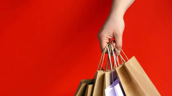 Hand with white paper bags and craft on bright red background. Season of sales or purchases as gift for holidays.Christmas shopping. Courier delivery of products in eco-friendly packaging. copy space