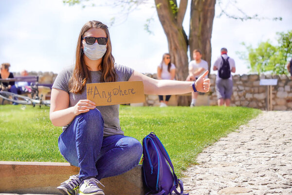 Girl in gray t-shirt, jeans, sunglasses and a medical mask with sign Anywhere is sitting by road, Humorous hitchhiking. Getting out of quarantine, traveling after coronavirus. Pandemic fear.