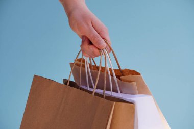 Beyaz kağıt torbalarla el ve parlak mavi arka planda el işi. Bayram hediyesi olarak Noel alışverişi. Noel alışverişi. Ürünlerin çevre dostu paketlemede kurye teslimatı. Boşluğu kopyala