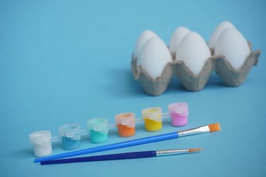 Mavi arka planda altı yumurta tepsisi ya da kutusu, çok renkli boya ve fırçalar. Paskalya 'da çocukların yaratıcılığına göre ayarlandı. Asgari paskalya konsepti. Sembol Mutlu Paskalya Düzlüğü