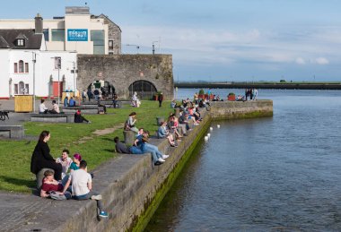 GALway, IRELAND - 21 Haziran 2017: Galway şehrindeki Corrib nehrinin kıyısında İspanyol Kemeri 'nin manzarasıyla sosyalleşen ve eğlenen insanlar.