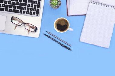 Blue office desk table with notebook, pen, glasses, cup of fresh espresso coffee and cactus. Top view with copy space, flat lay