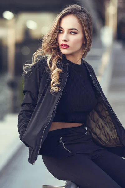 Mooi jong meisje dragen zwarte jas zitten in de straat. — Stockfoto