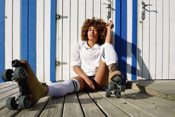 Joven mujer negra en patines sentada cerca de una cabaña de playa . — Foto de Stock