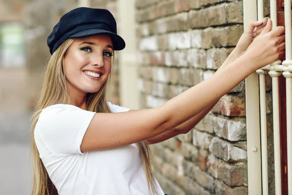 Mujer rubia joven con gorra sonriendo cerca de una pared de ladrillo . — Foto de Stock