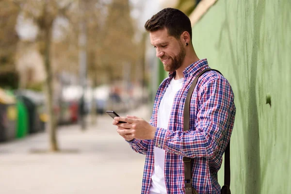 微笑的年轻人看着他的智能手机在城市背景。我 — 图库照片
