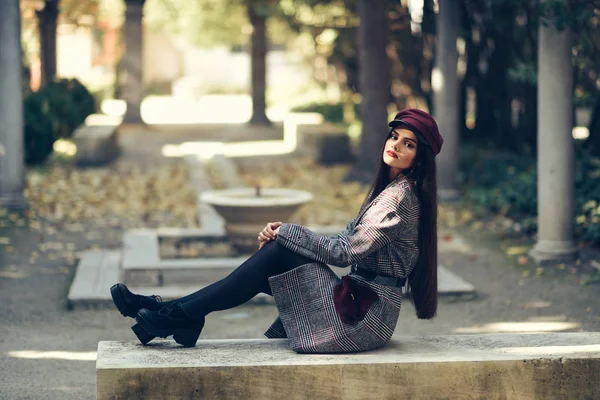 Joven hermosa chica con abrigo de invierno y gorra sentado en un banco en el parque urbano . — Foto de Stock