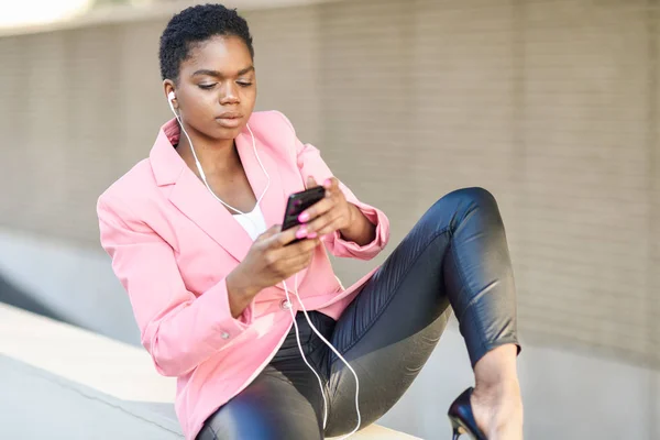 Empresaria negra sentada al aire libre usando smartphone con auriculares —  Fotos de Stock