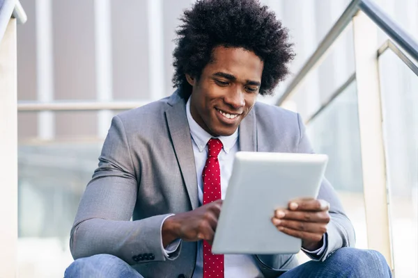 Hombre de negocios negro usando una tableta digital sentada cerca de un edificio de oficinas. —  Fotos de Stock