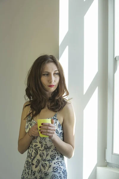 Sweet girl drinks coffee near a window in broad daylight. It looks like she\'s waiting for something and is thoughtful