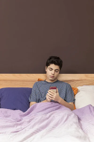 Boy in the bed with phone, he lose just time and maybe he chatting or he looks to something. Front position.
