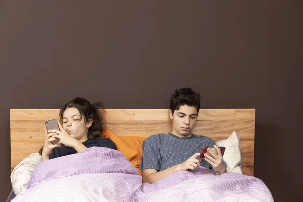 A young girl and a young boy together in the double bed. Even if they are together everyone is alone with his phone.
