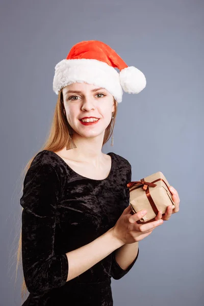 Jovem caucasiana senhora em um chapéu de Natal vermelho e vestido preto está dando uma caixa de presente para você para mim em fundo cinza. isolado — Fotografia de Stock