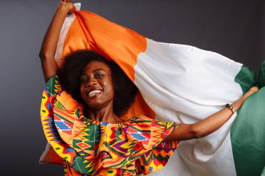 Ulusal giysiler içinde mutlu bir Afrikalı kadın gülümsüyor ve Fildişi Sahili bayrağıyla poz veriyor.