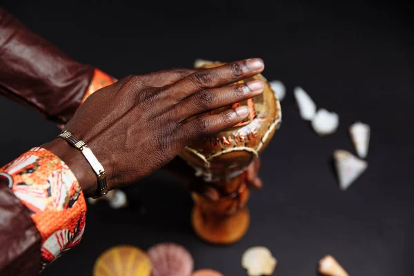 Las manos de un hombre africano con ropa nacional sostiene tambor djembe rodeado de conchas marinas — Foto de Stock