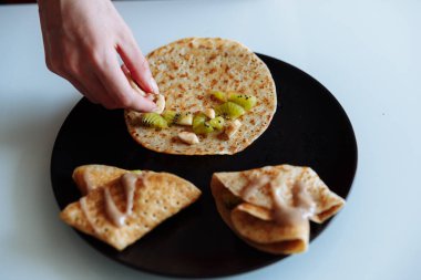 Kadının eli krepleri meyveli kivi ve muzla süslüyor. Siyah tabak, beyaz masa..