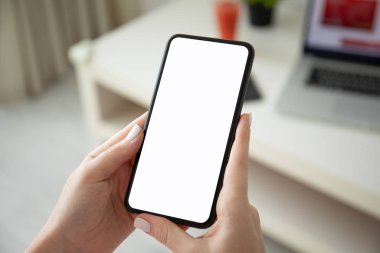 Kadın elleri, odada dizüstü bilgisayarı olan bir masanın üzerinde telefonu tek başına tutuyor.