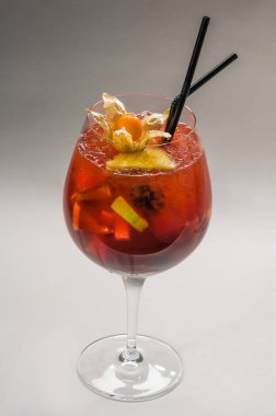 Şeffaf cam kadehte kokteyl, fizsalis, portakal ve çilek parçaları, buz parçaları ve hafif arka planda kokteyl için siyah pipetler.