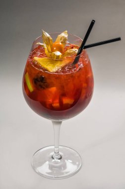 Şeffaf cam kadehte kokteyl, fizsalis, portakal ve çilek parçaları, buz parçaları ve hafif arka planda kokteyl için siyah pipetler.