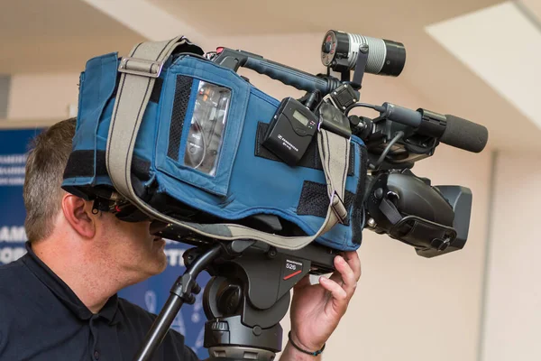 An operator with a professional video camera under a tripod against the background of the room.