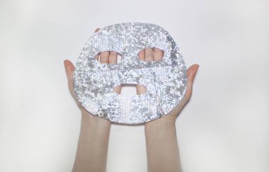 Kadın eller yüz için bir kumaş holografik gümüş çarşaf maskesi tutuyor. Beyaz bir arka planda. Yukarıdan görüntüle. 