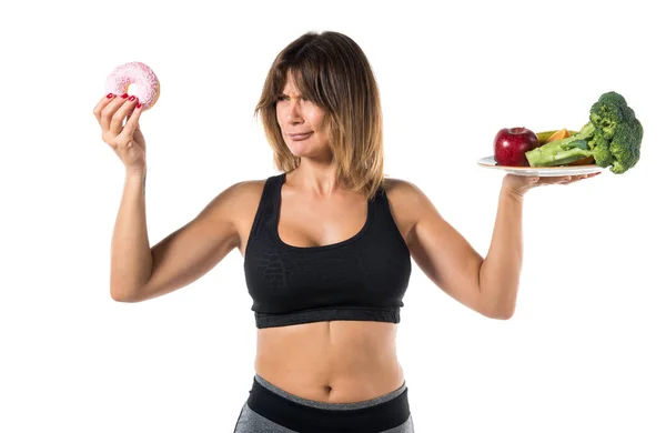 Sport woman holding fruits and vegetables in one hand and donut in other hand — Stock Photo, Image