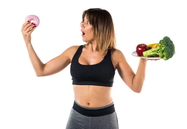Sport woman holding fruits and vegetables in one hand and donut in other hand — Stock Photo, Image